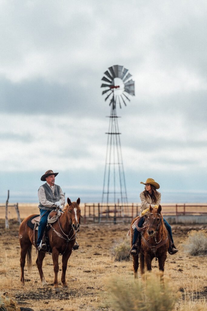 Man and woman sitting on horses in a field with windmill behind them.