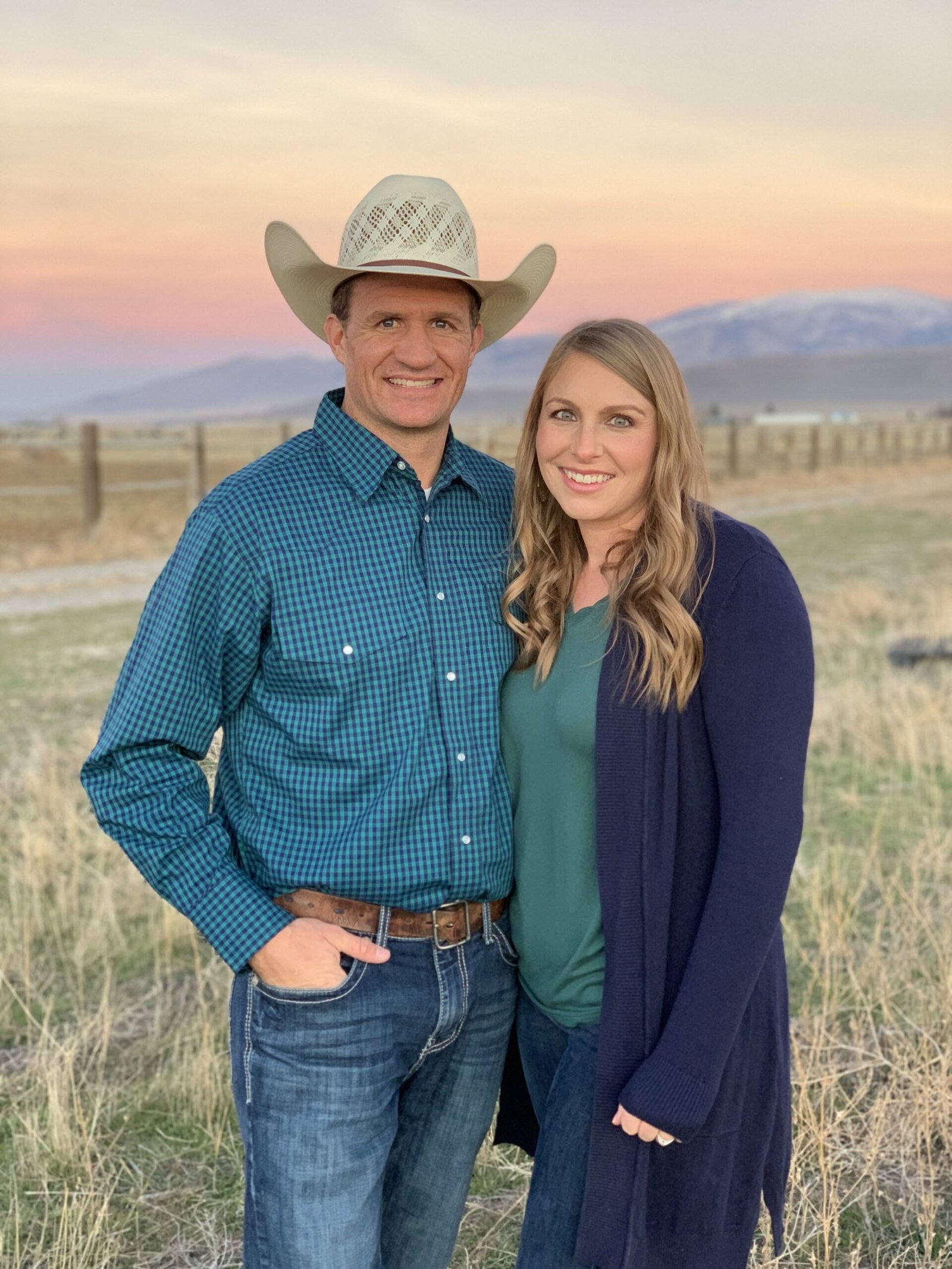 Man and woman standing in field with mountains and a beautiful sunset behind them. Click to view Hat Brand Beef's farm profile.